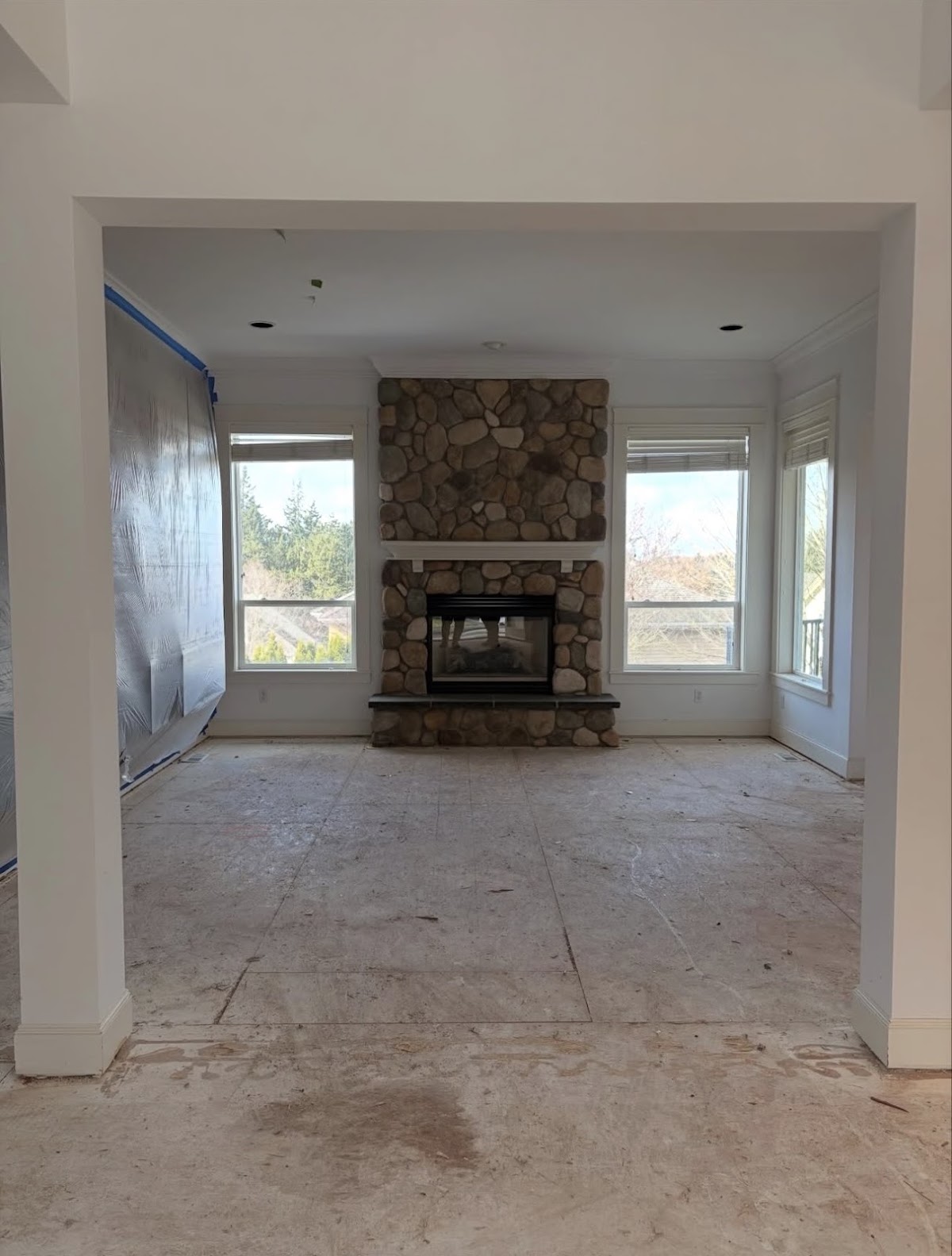 The same living room, emptied and swept down to bare subfloor, the stone fireplace and windows unchanged.